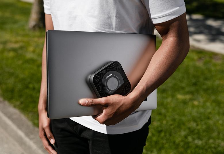 An image of a person carrying the Nighthawk M1 4G LTE Mobile Router (MR1100) in a park while using a laptop. The router provides reliable internet connectivity on the go, allowing users to work or browse the internet from outdoor locations.
